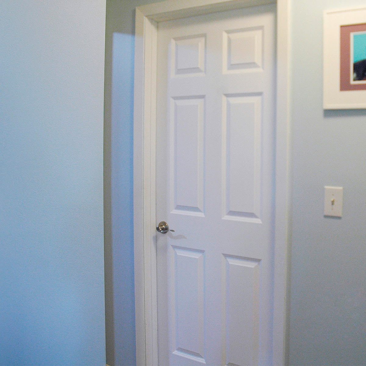 A white six-panel door stands ajar in a room with light blue walls, framed artwork nearby, suggesting an inviting, simple interior space.