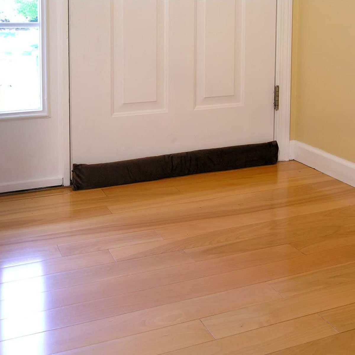 A dark draft stopper rests against the bottom of a closed door, preventing air movement in a room with polished wooden floors and yellow wal