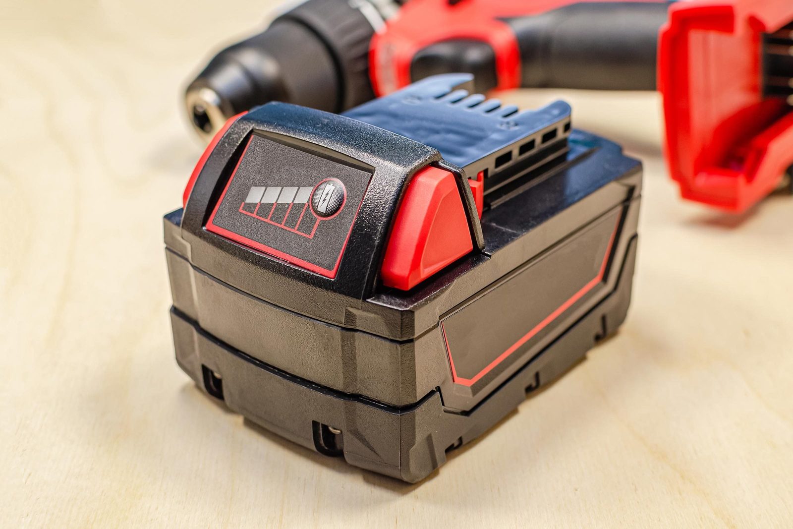 A battery sits on a wooden surface, displaying charge indicators, while an electric drill with a red and black design is partially visible n