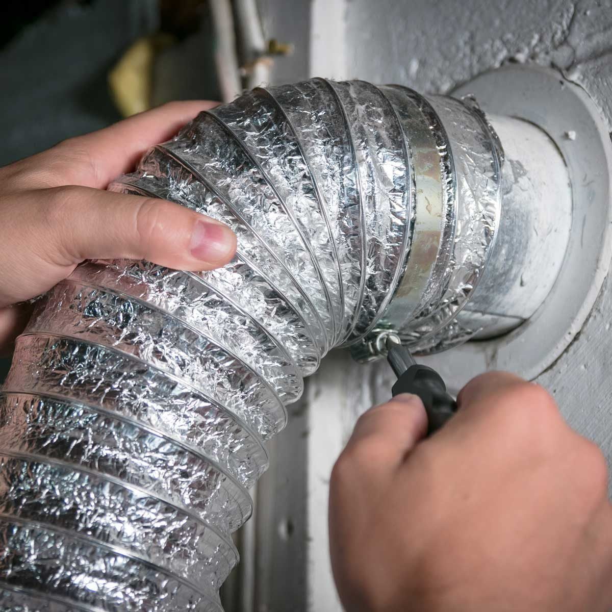 A metallic duct is being secured with a tool, while hands grip the duct and the surrounding wall is painted in light tones.