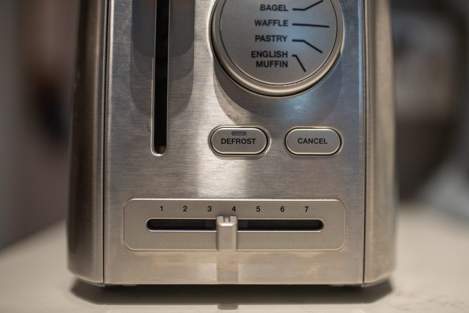 A stainless steel toaster sits on a countertop, featuring settings for various bread types and a sliding timer dial from 1 to 7.