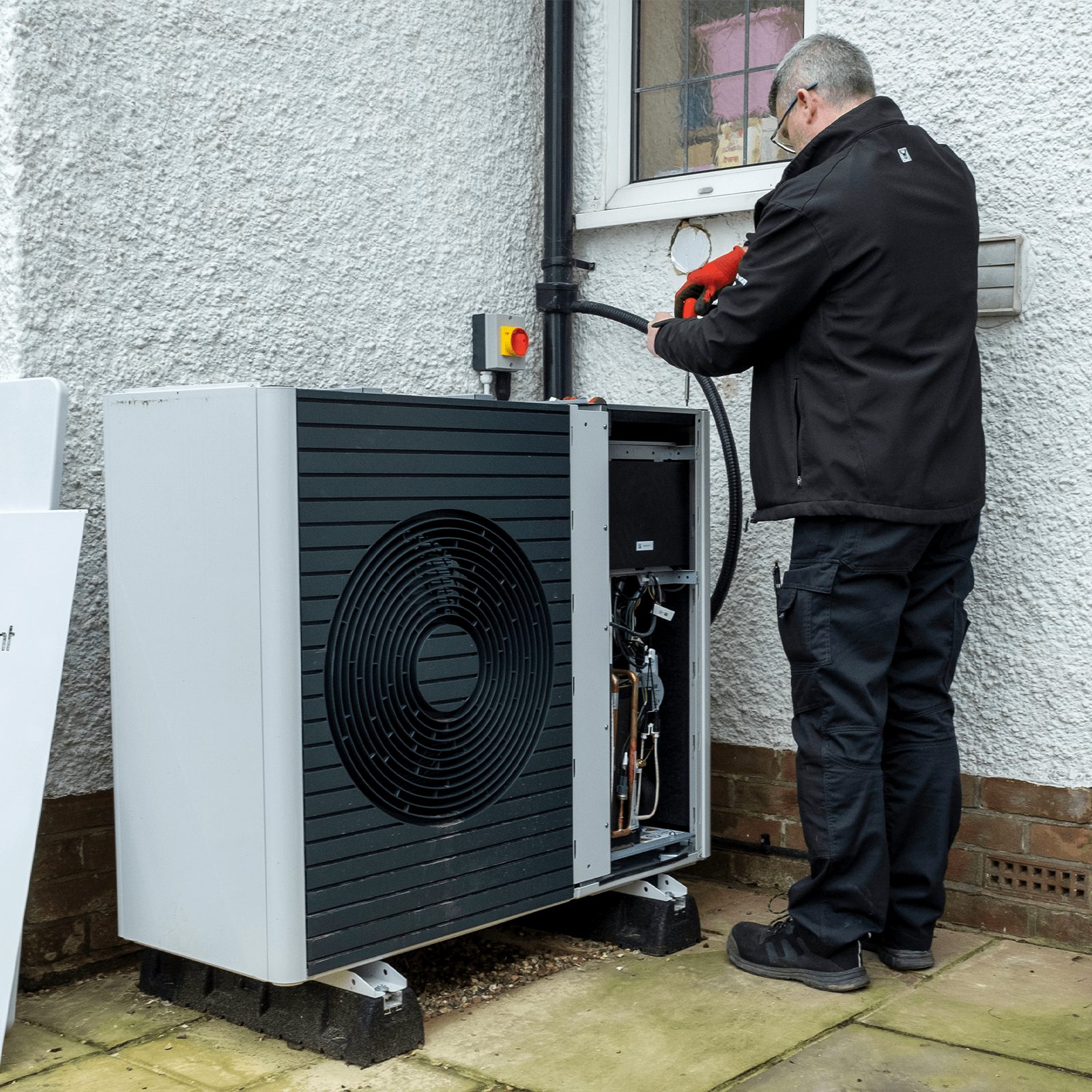 A technician connects a hose to an outdoor heat pump beside a textured white wall, surrounded by concrete paving and a small garden.