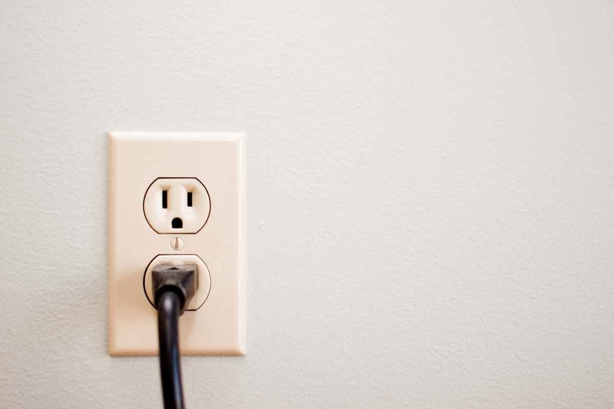 An electrical outlet is mounted on a light-colored wall, with a black plug inserted into it. The surrounding area is simple and uncluttered.