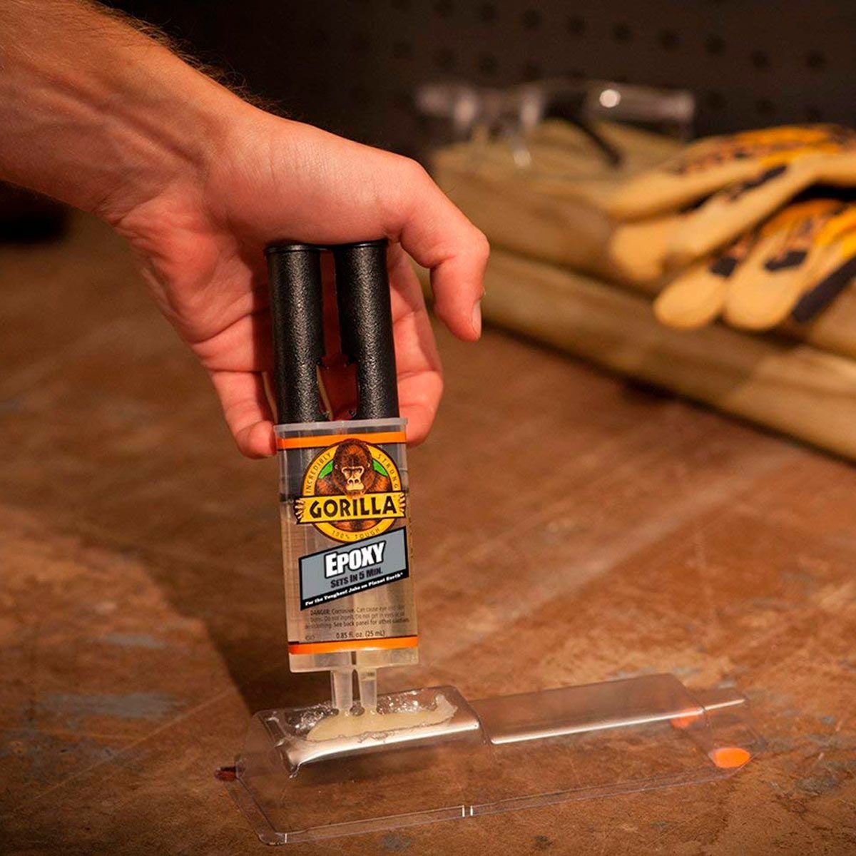 A hand squeezes a Gorilla epoxy dispenser, dispensing adhesive onto a transparent surface, with work gloves and tools visible in the backgro