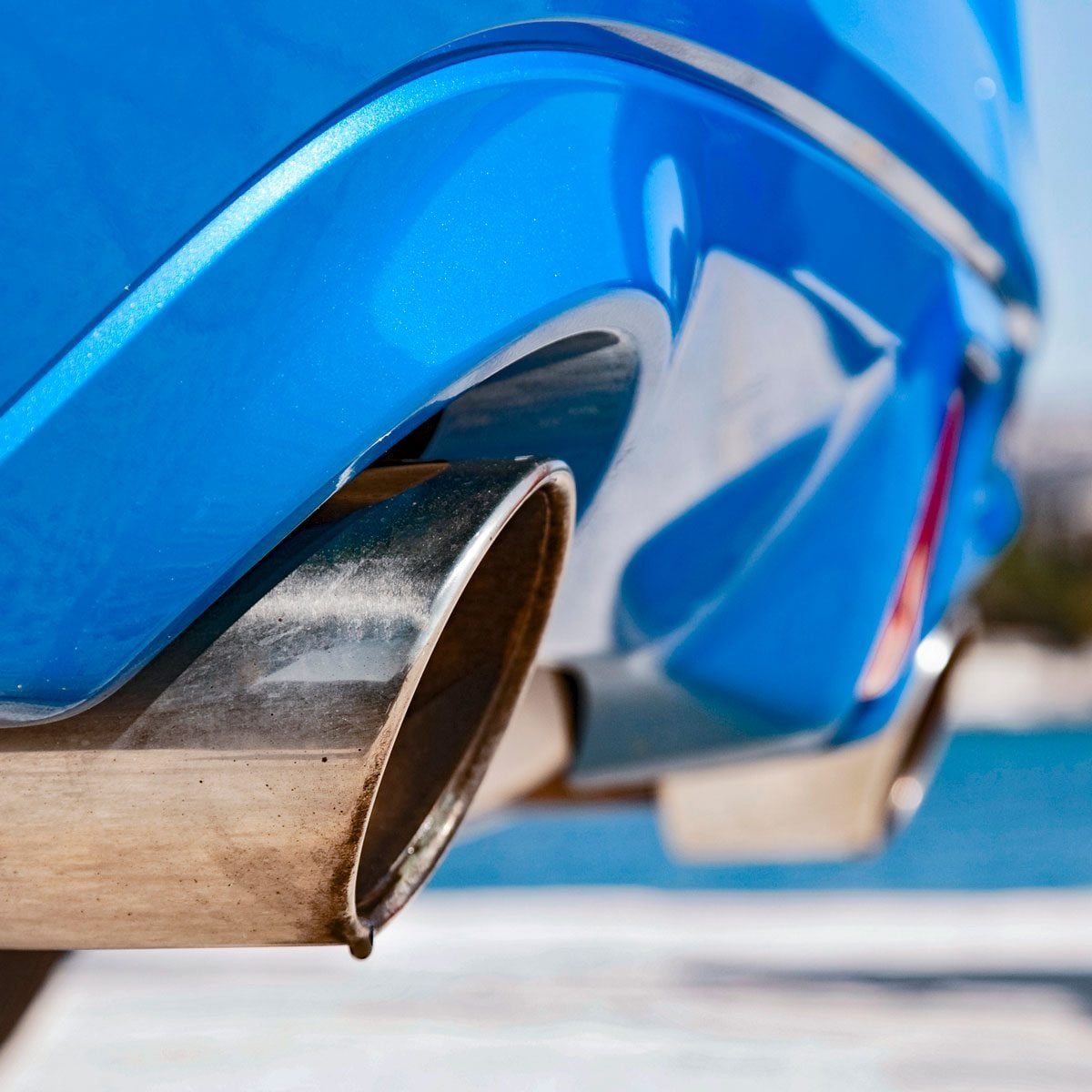 A shiny, metallic exhaust pipe releases exhaust gases, positioned below a bright blue car's body, with a blurred background of a blue sky an