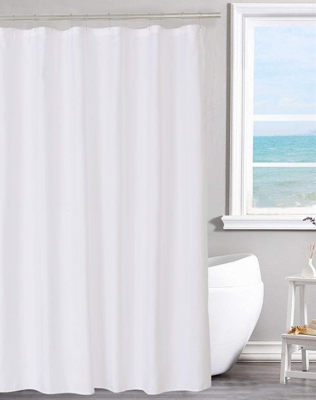 A white shower curtain hangs in a bathroom, separating the bathing area from a toilet and a window revealing a view of the ocean.