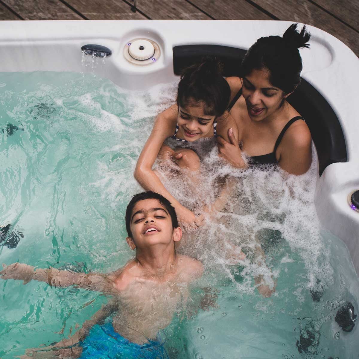 Three people play in a hot tub, splashing water joyfully. Two children are engaged with a woman, laughing and enjoying the moment on a woode