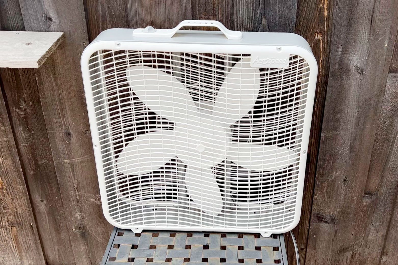 A white box fan stands upright on a metal grid surface, surrounded by brown wooden walls, ready to circulate air.
