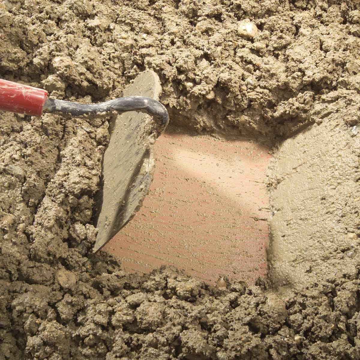 A trowel scrapes against a smooth surface exposed in wet, muddy soil, surrounded by loose dirt and debris, indicating a construction or exca