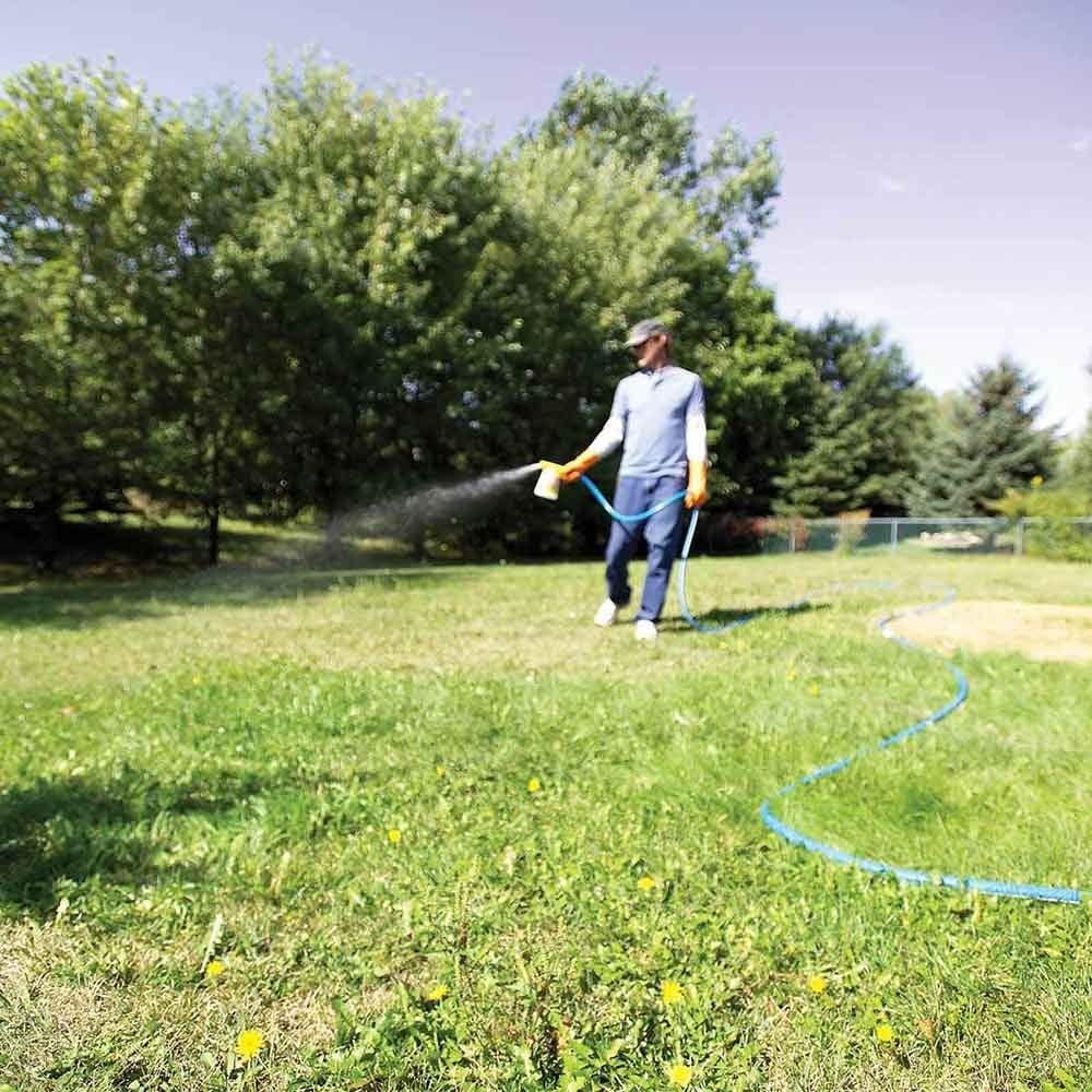 A person sprays liquid on grass in a garden, surrounded by trees and a blue hose laying on the ground under a clear sky.