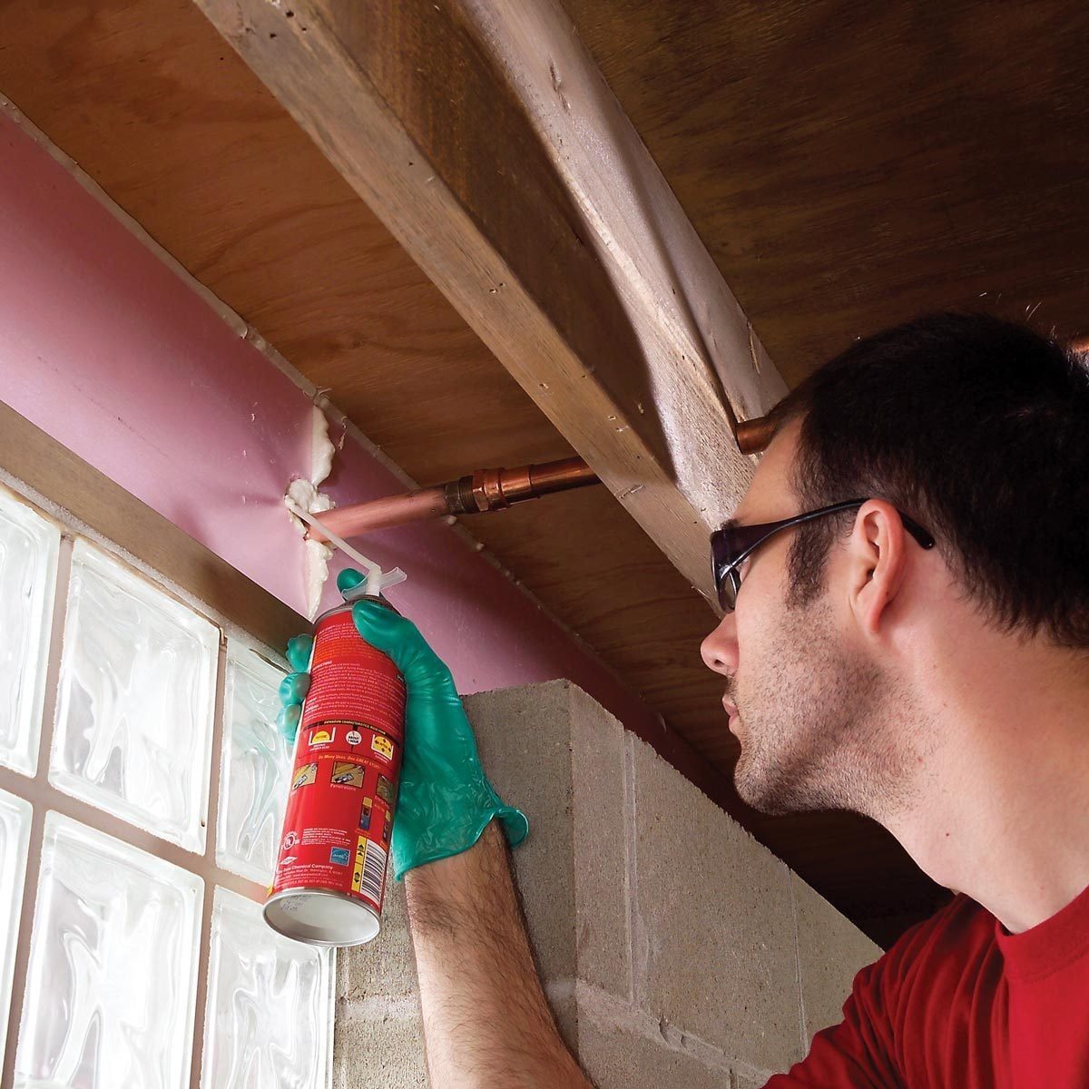 A person wearing gloves and glasses applies sealant from a spray can to a gap near plumbing in a construction area with concrete walls.