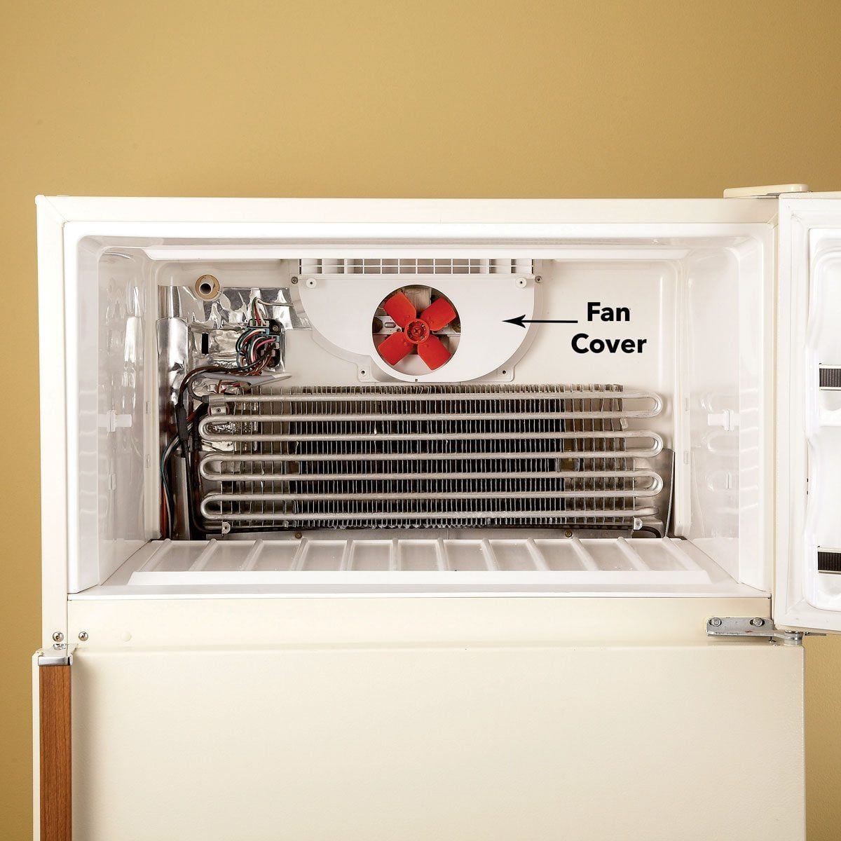 An open refrigerator interior features a red fan behind a cover and metal coils for cooling, set against a beige wall.