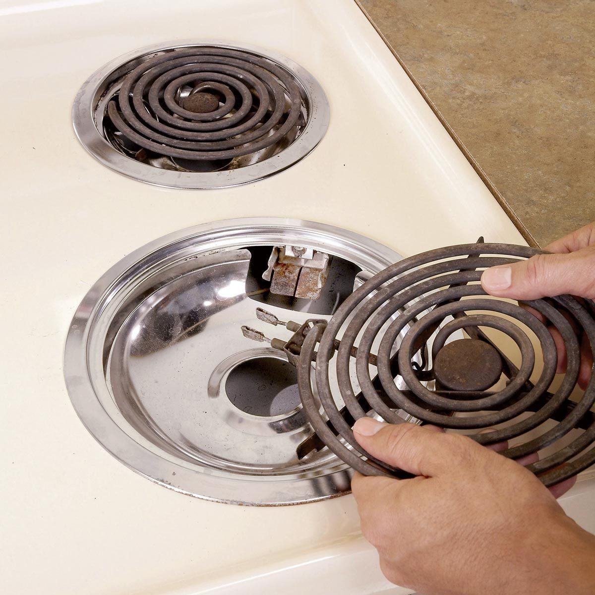 A person is removing a metal stove coil from a stovetop, revealing a shiny circular area with electrical connections, on a light-colored cou
