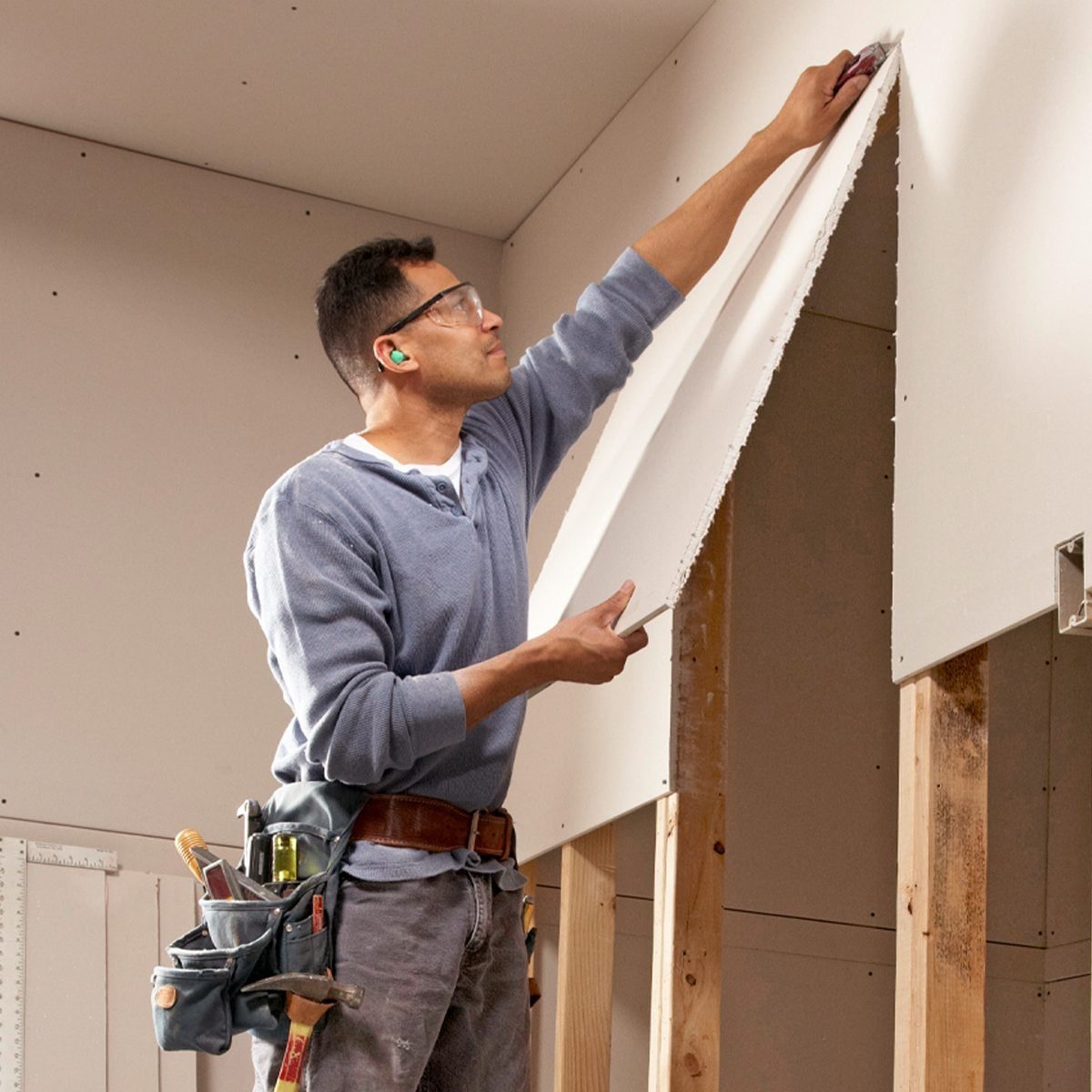 A man applies drywall to a wall, positioned in a partially constructed room, wearing safety glasses and ear protection, with tools in a belt