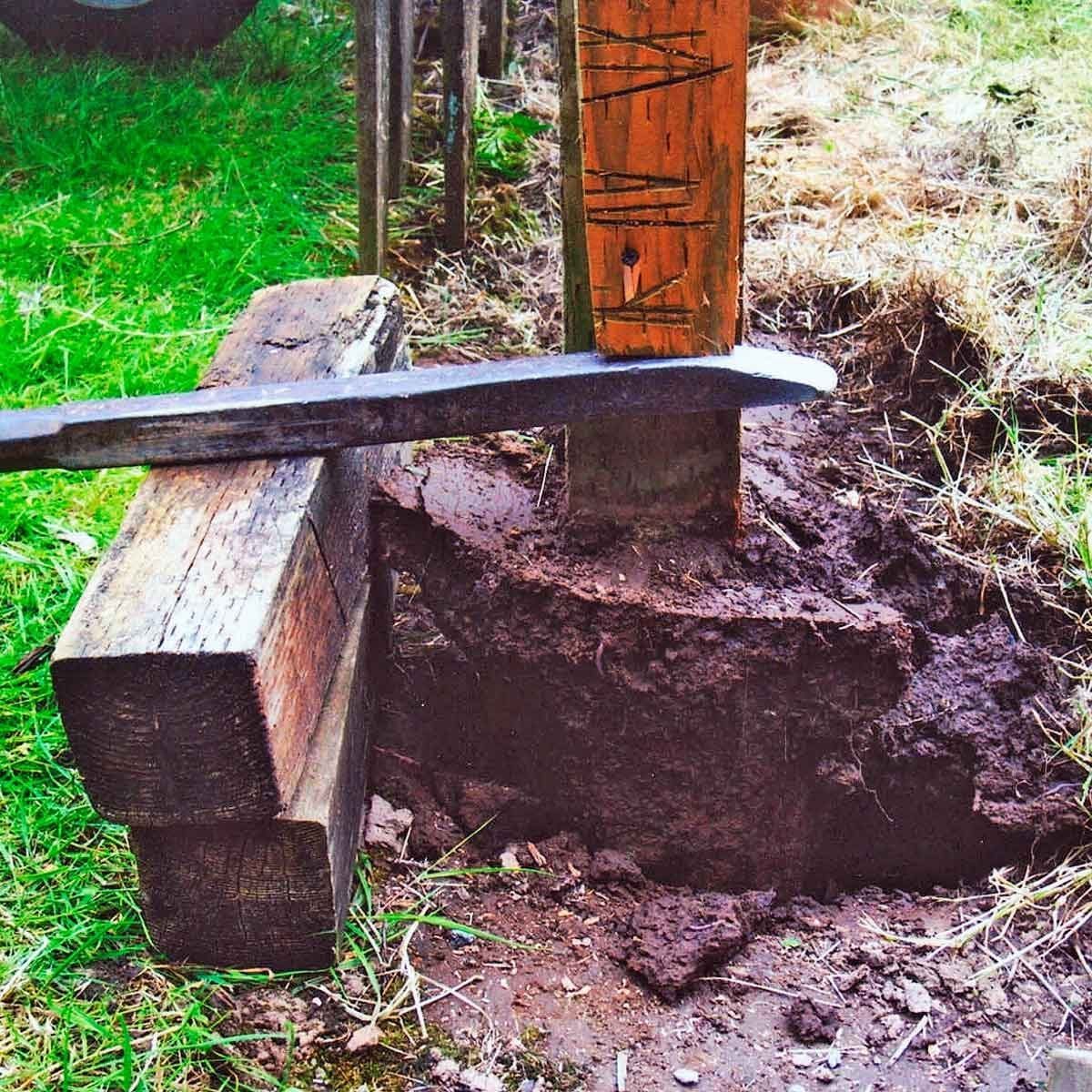 A metal tool is positioned against a wooden post, while soil is being excavated around it in a grassy outdoor area.