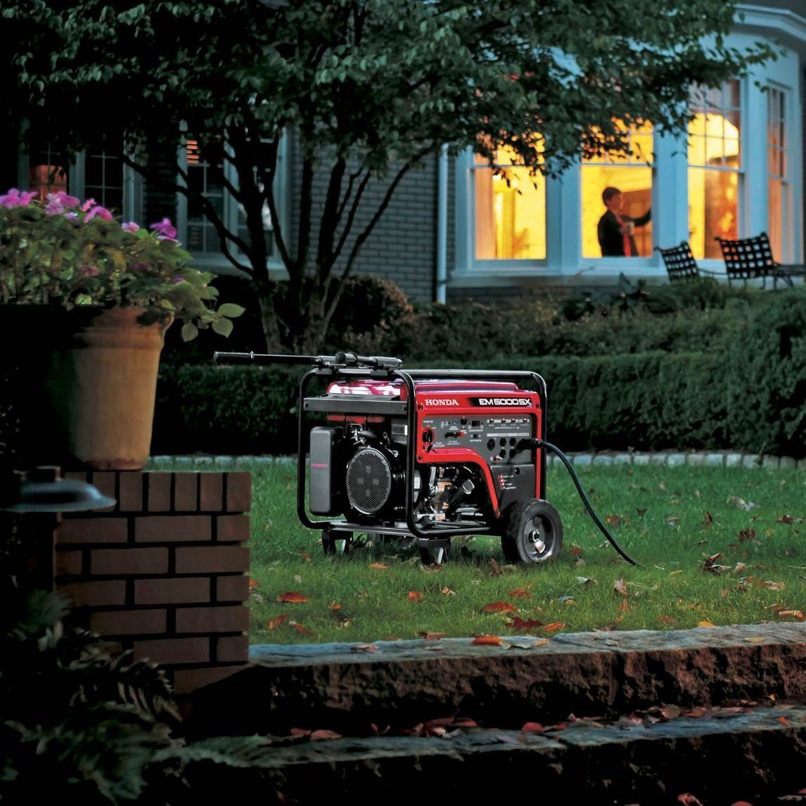 A portable generator is placed on a lawn in the foreground, with a large house in the background displaying illuminated windows. Inside the house, two people are visible through