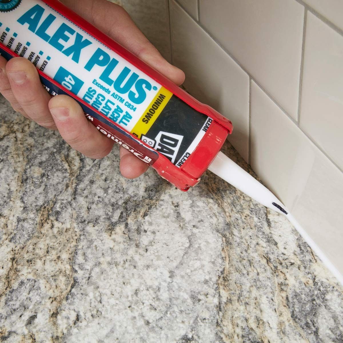 A hand holds a caulking gun, applying sealant against a tiled wall, with a granite countertop beneath. The setting appears to be a kitchen o