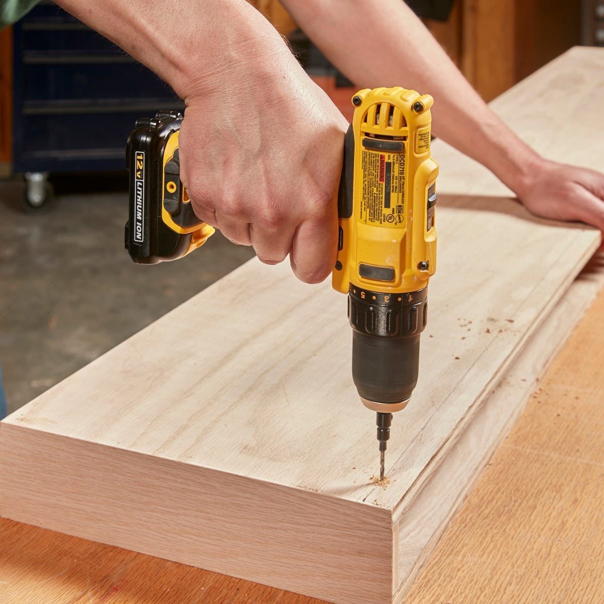 A person uses a yellow power drill to bore a hole into a wooden plank on a work surface in a workshop.