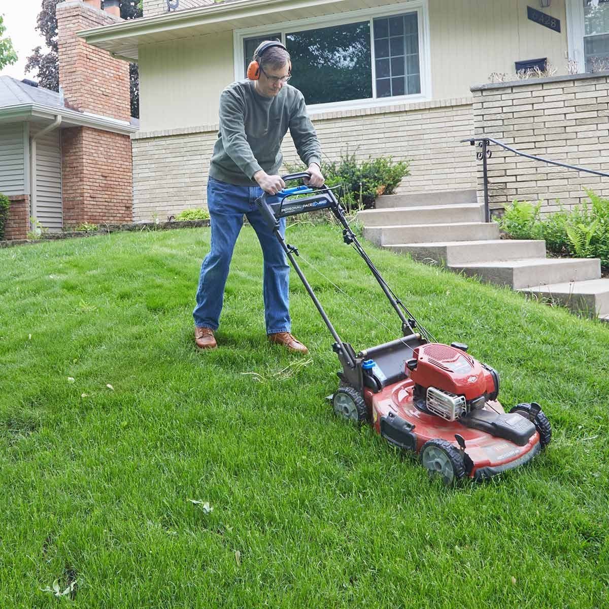A man mows a green lawn using a gas-powered mower in a residential yard, surrounded by steps leading to a house with a brick facade.