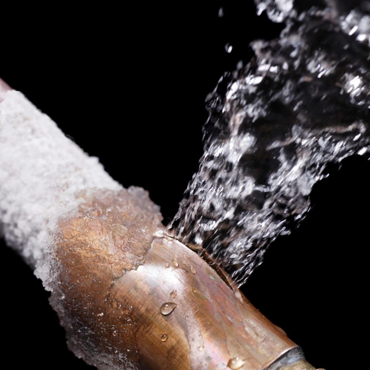 A copper pipe leaks water, spraying forcefully from a cracked joint, surrounded by dark space with droplets glistening on the surface.