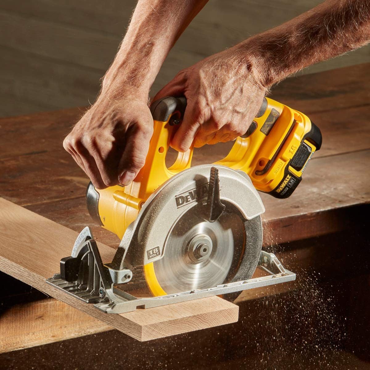 A circular saw, held with two hands, cuts through a wooden plank, sending sawdust flying in a workshop environment.