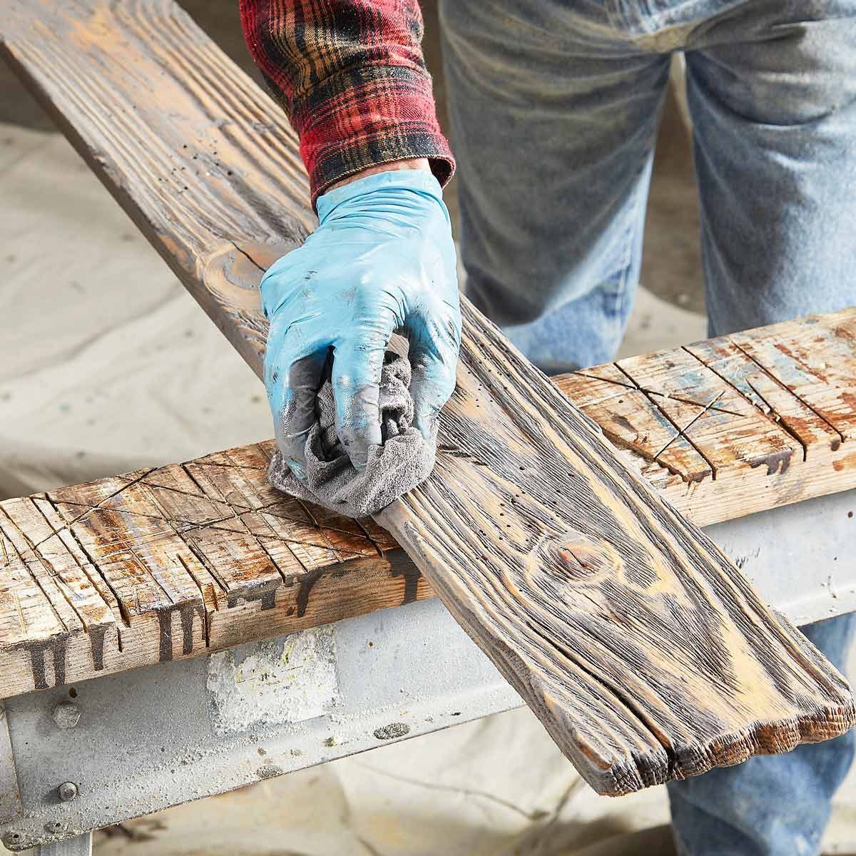 A gloved hand wipes a wooden plank with a cloth, while standing in a workshop environment with sawdust and tools visible in the background.