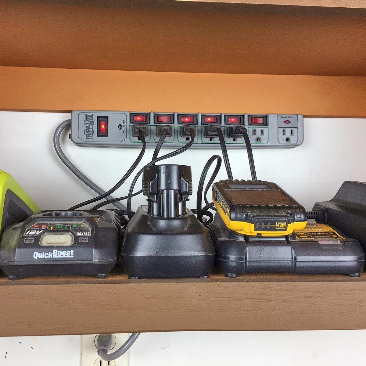 A power strip with multiple red indicator lights is mounted on a wall, while several battery chargers for tools rest on a shelf below it.