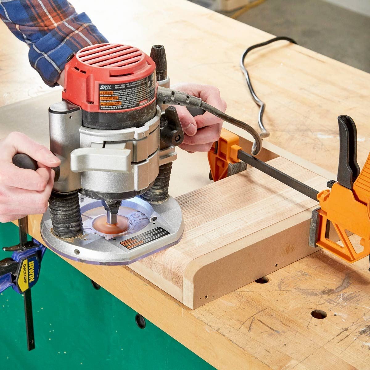 A router is being used to shape the edge of a wooden piece, securely held by a clamp on a workbench surrounded by tools and sawdust.