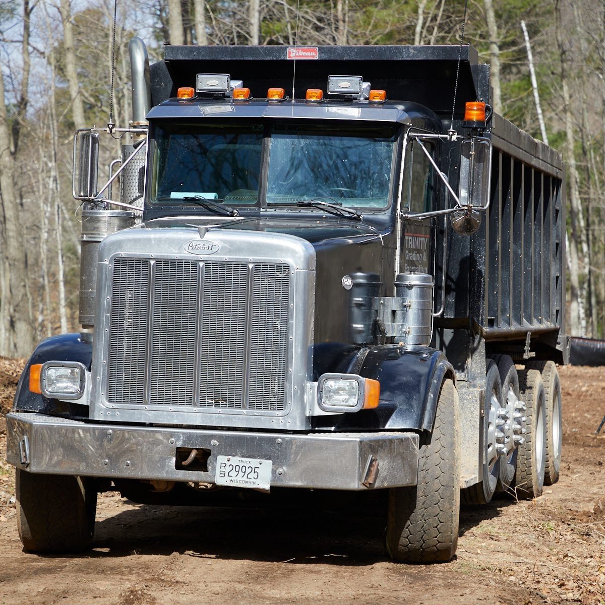 A black dump truck is parked on a dirt site surrounded by trees, with its engine running and lights activated, ready for loading work.