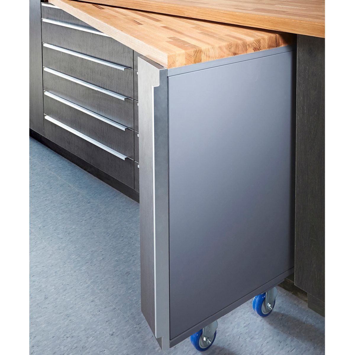 A metal rolling cabinet with a wooden countertop is positioned against a gray floor, featuring drawers above and a smooth, gray surface on t