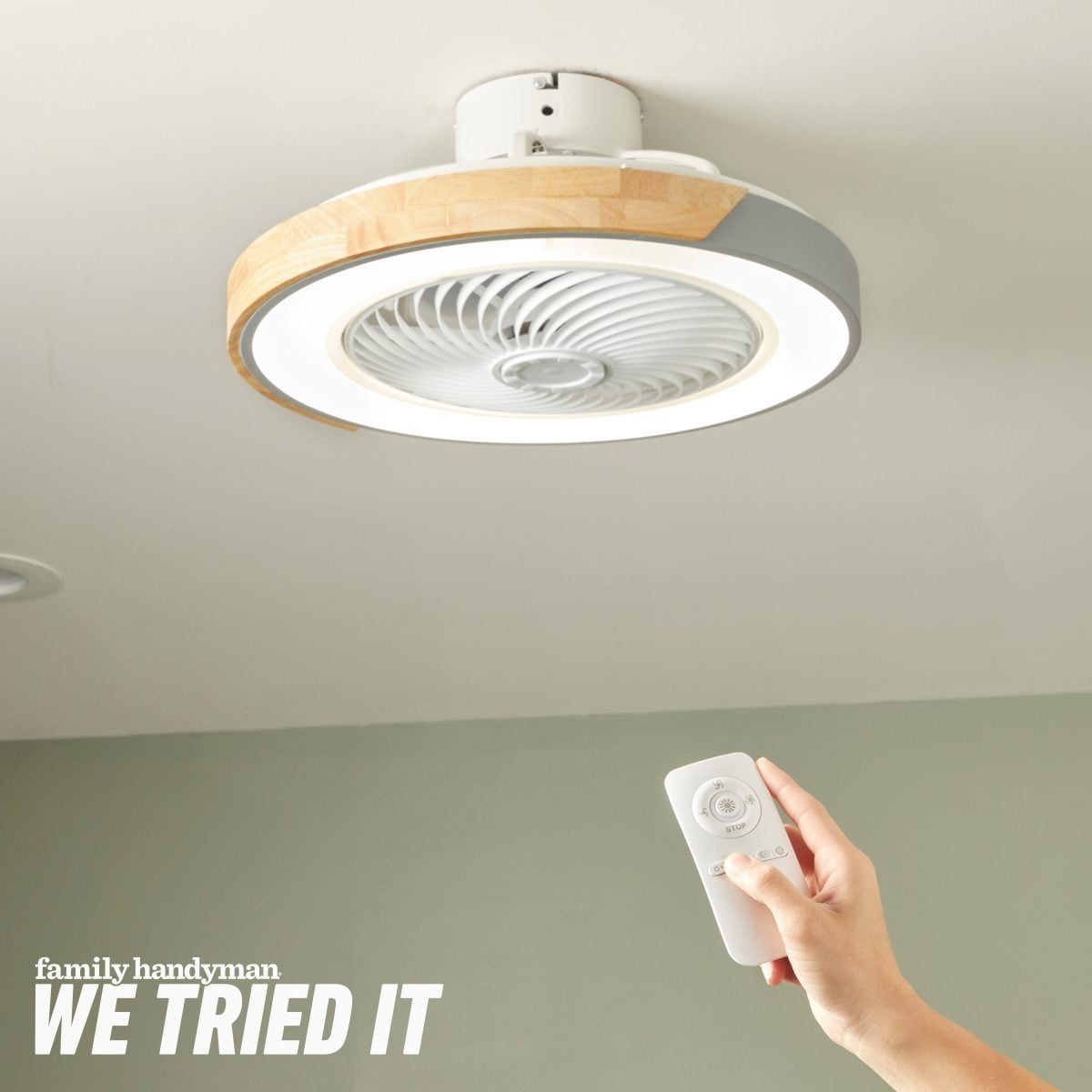 A ceiling fan with a light is mounted overhead, while a hand holds a remote control, likely adjusting the fan's settings in a neutral-colore