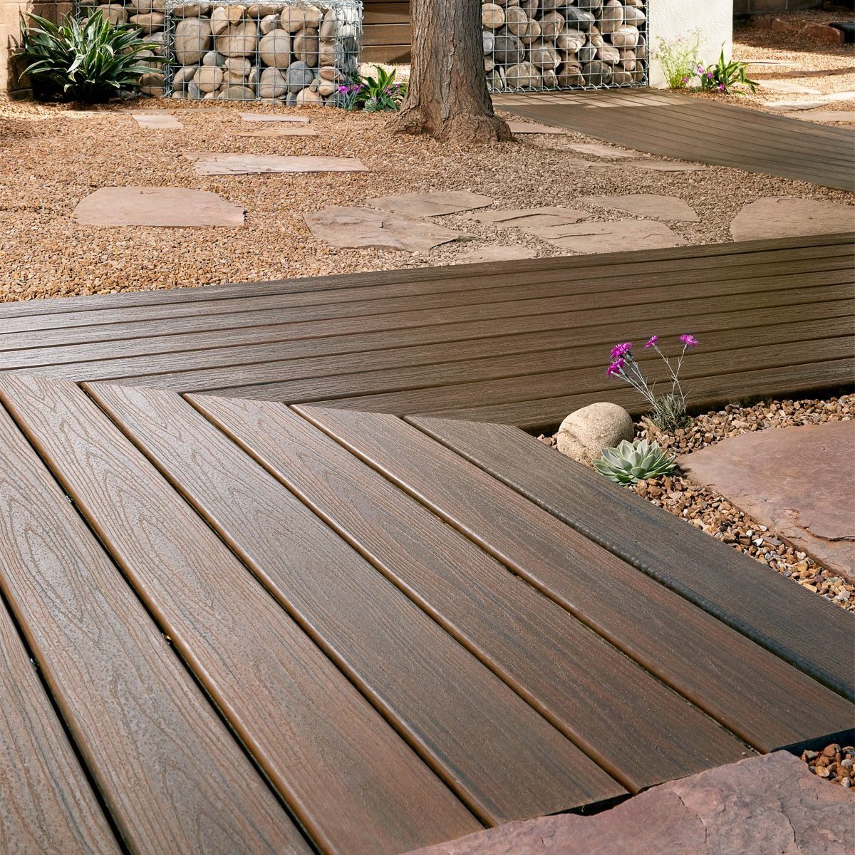 Wooden decking boards create a pathway, intersecting in a zigzag pattern across a gravel landscape, surrounded by stone accents and small pl