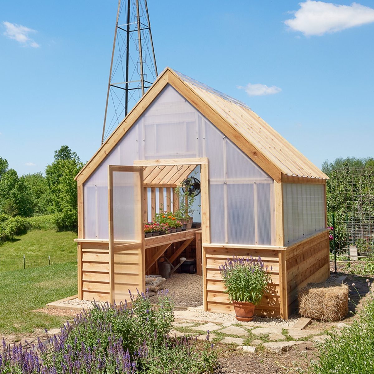 A wooden greenhouse with an open door displays potted flowers inside, surrounded by greenery, gravel paths, and lavender plants under a clea