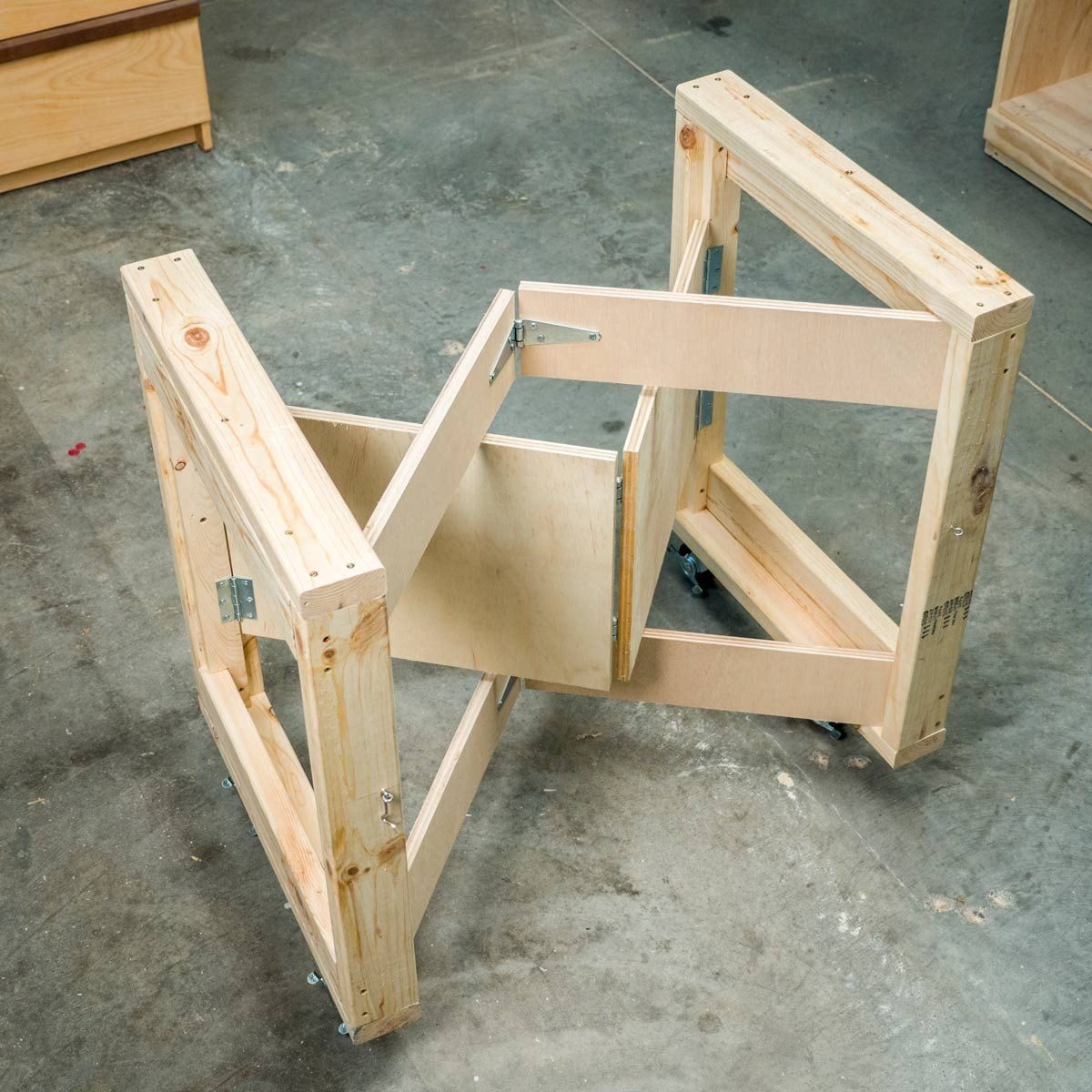 A wooden table frame is partially collapsed, showing open compartments. It rests on a concrete floor in a workshop environment.