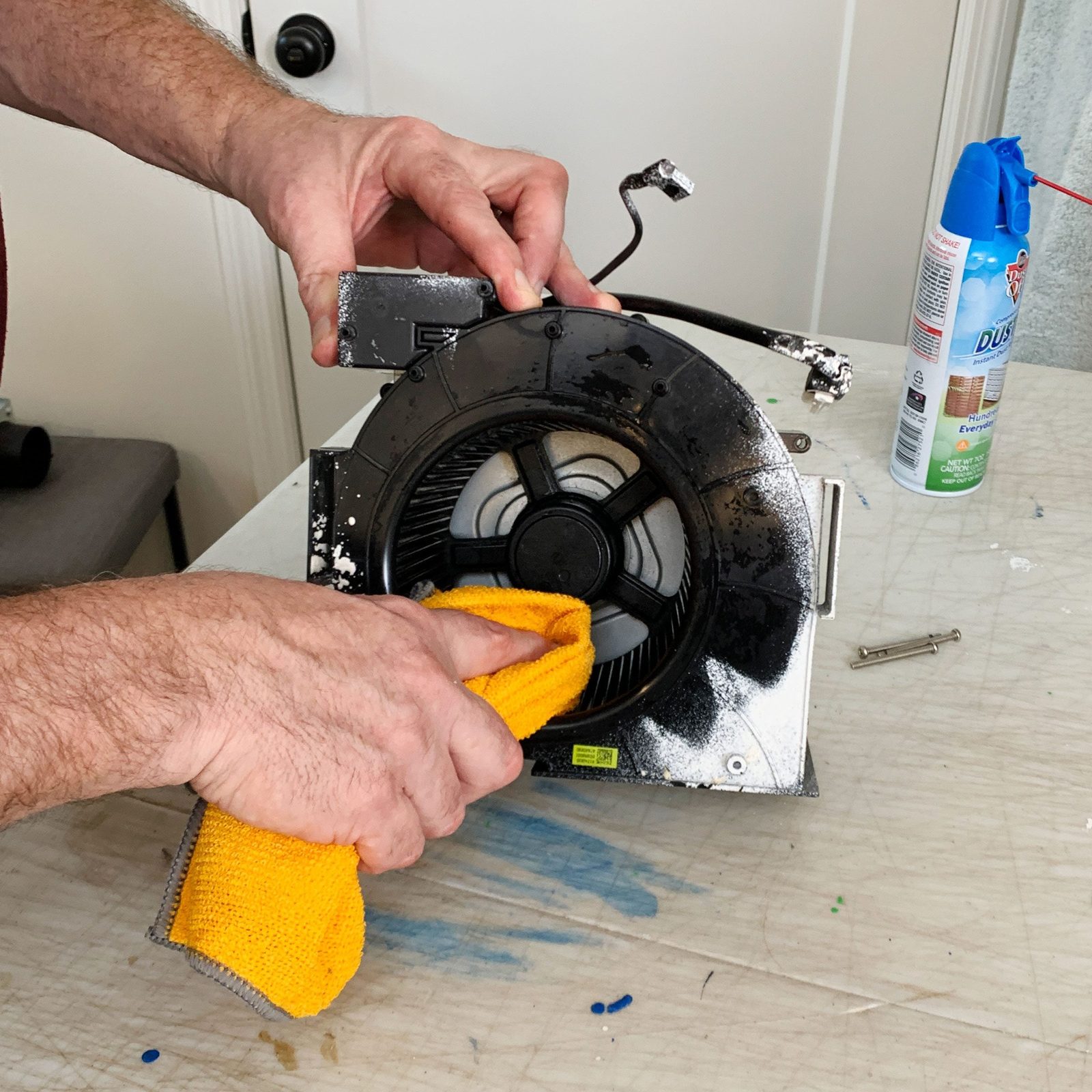 A person cleans a black fan unit with a yellow cloth on a work surface, surrounded by cleaning spray and small tools.