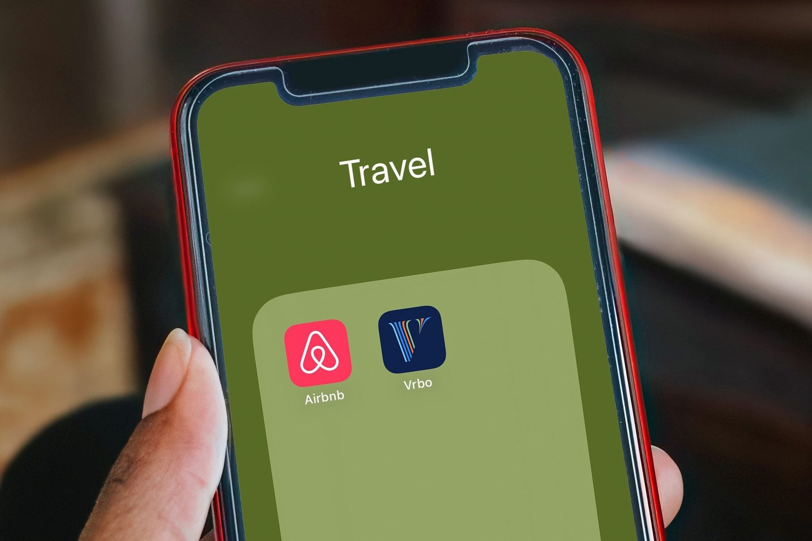 A hand holds a smartphone displaying a green screen with a folder labeled "Travel," containing icons for Airbnb and Vrbo applications.