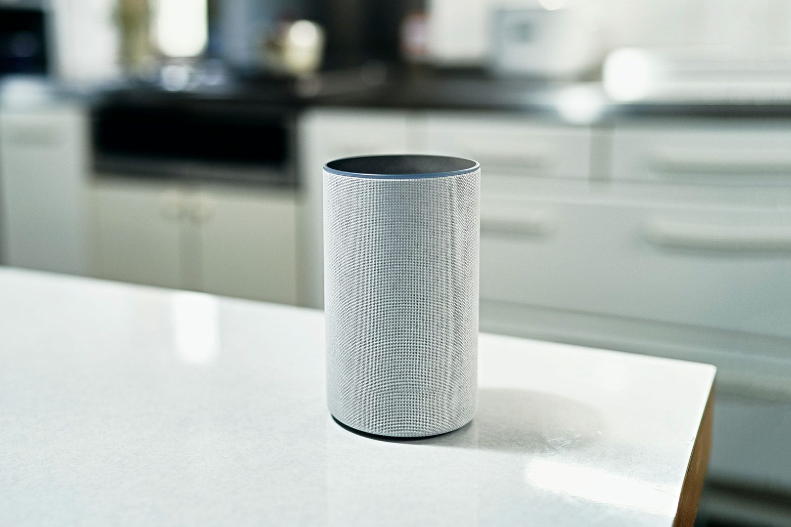 A cylindrical smart speaker stands on a kitchen countertop, surrounded by blurred cabinets and appliances, suggesting a modern home environm
