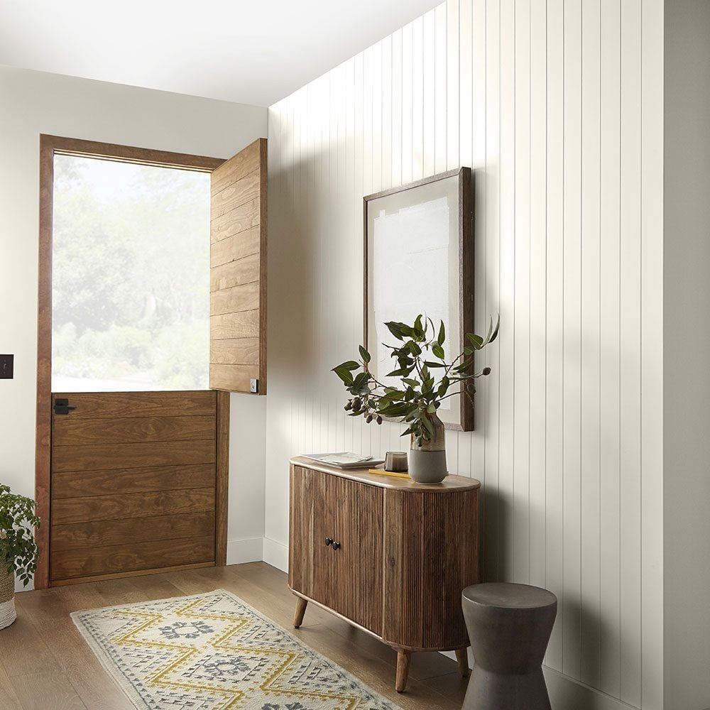 A wooden door opens, revealing a bright entryway with a cabinet, a plant, and a decorative rug on the floor, accented by vertical paneling.
