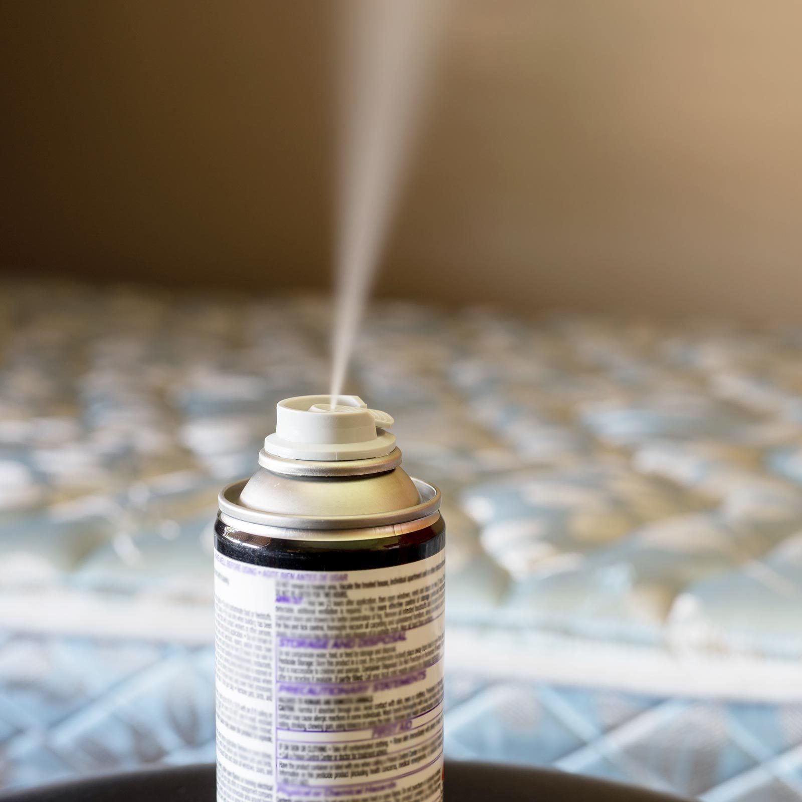 A spray can releases a fine mist, positioned on a table in front of a patterned mattress, indicating a cleaning or freshening action.