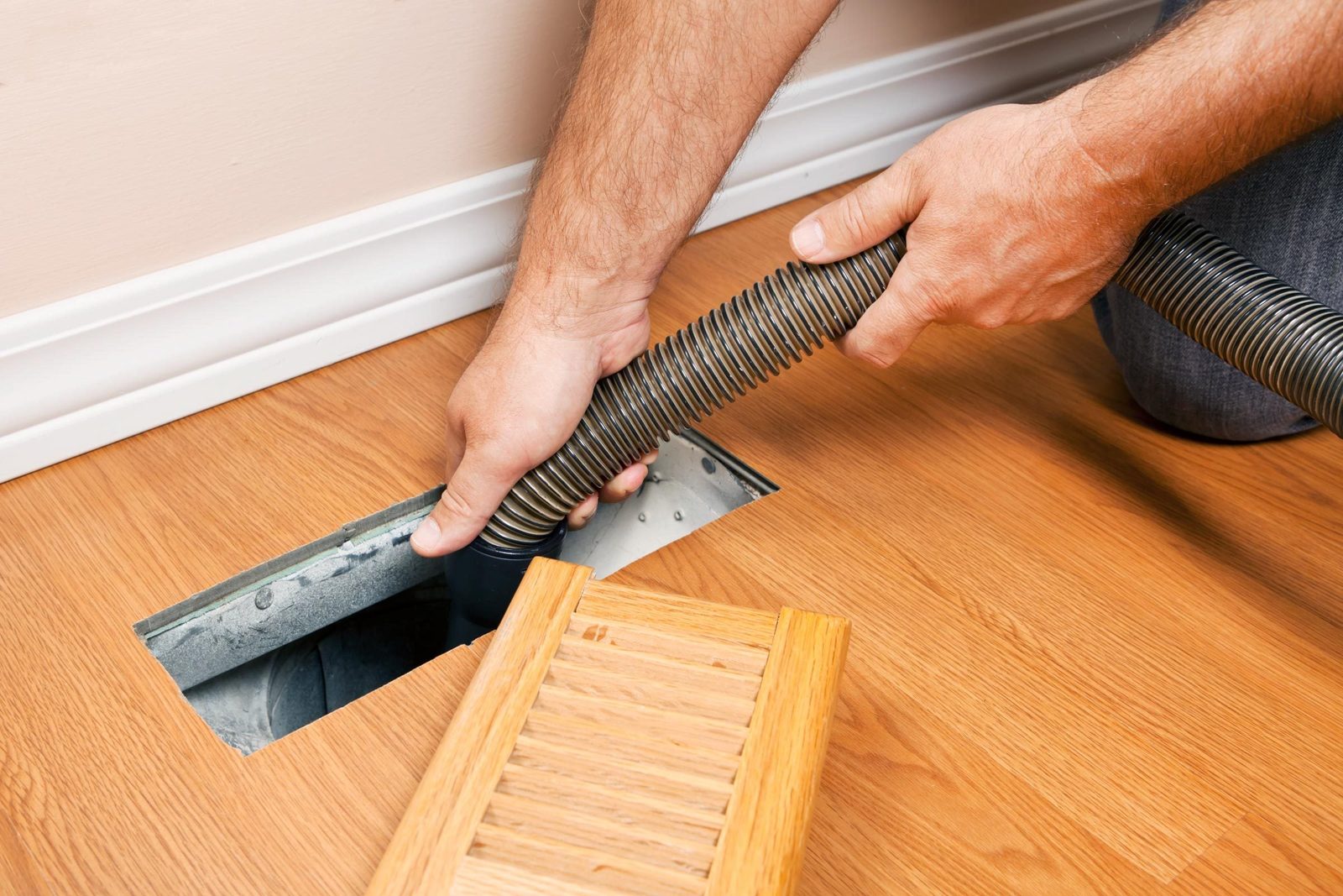 A person is using a vacuum hose to clean dust from a floor vent in a room with wooden flooring and a light-colored wall.