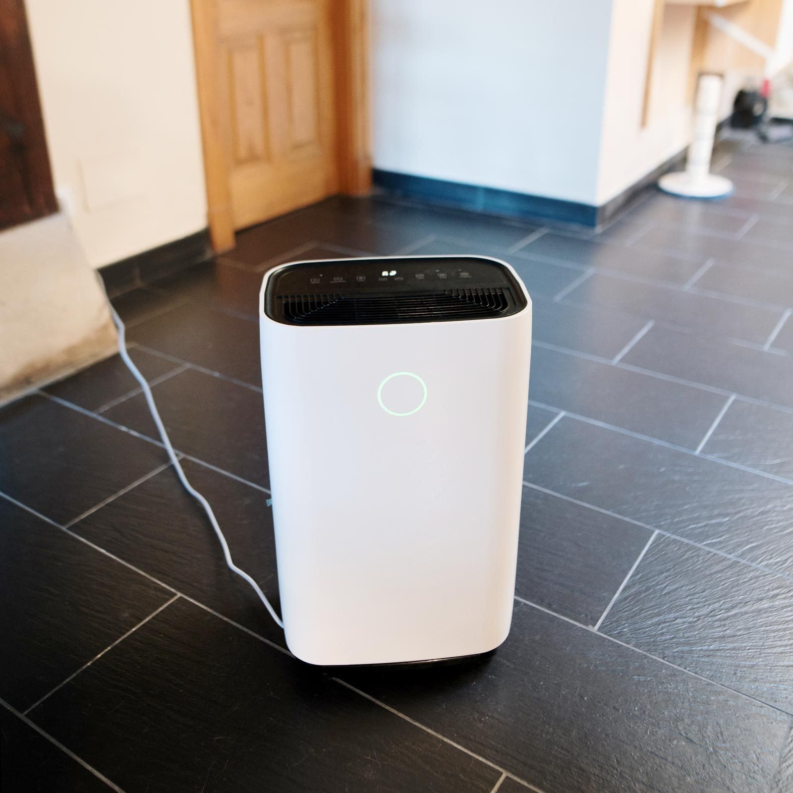 A white air purifier stands on a dark tiled floor, plugged in, with a faintly glowing indicator light on its front, in a room with wooden ac