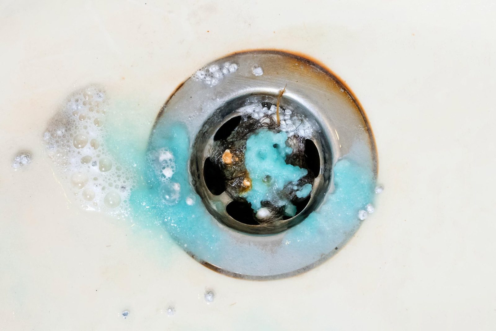 A drain with a metallic cover shows blue foam and bubbles surrounding debris, indicating stagnant water in a white sink.