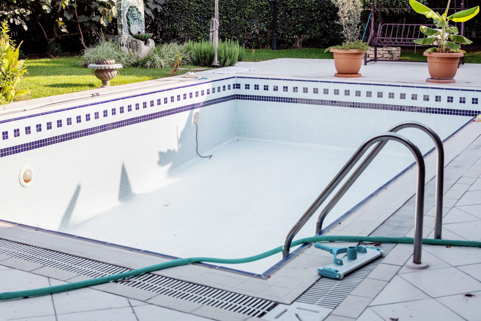 An empty swimming pool is being cleaned with a vacuum, surrounded by a tiled deck, green plants, and a stone ornament in a garden setting.