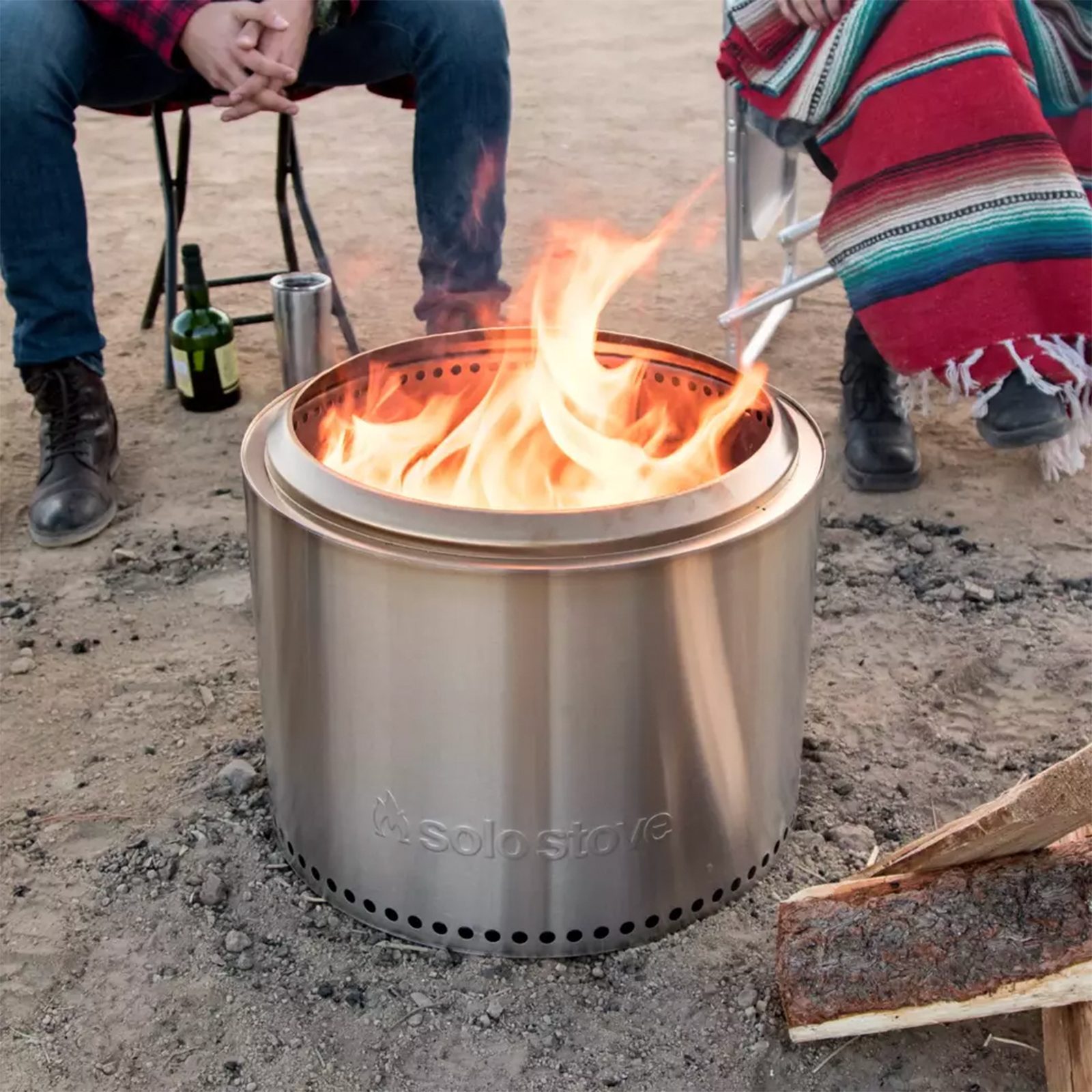 A stainless steel fire pit burns brightly. People sit nearby on a dirt surface, one wrapped in a colorful blanket, with drinks beside them.