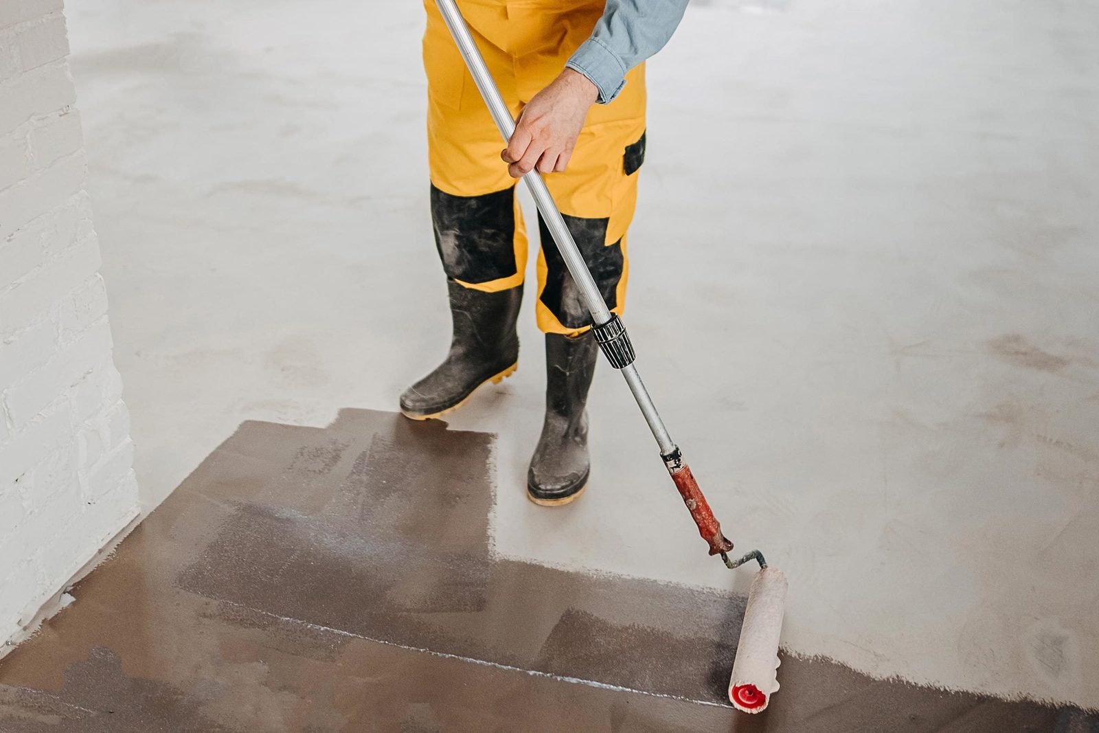 A person in yellow overalls rolls paint onto a freshly applied brown surface, standing in a spacious, unfinished room with a light-colored f