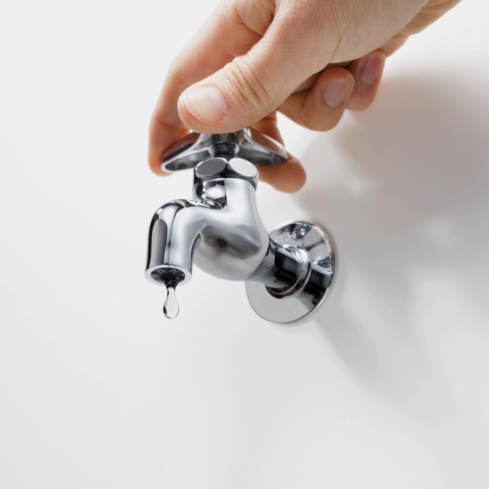 A hand turns a chrome faucet, and a single droplet of water hangs from the spout against a plain white wall.