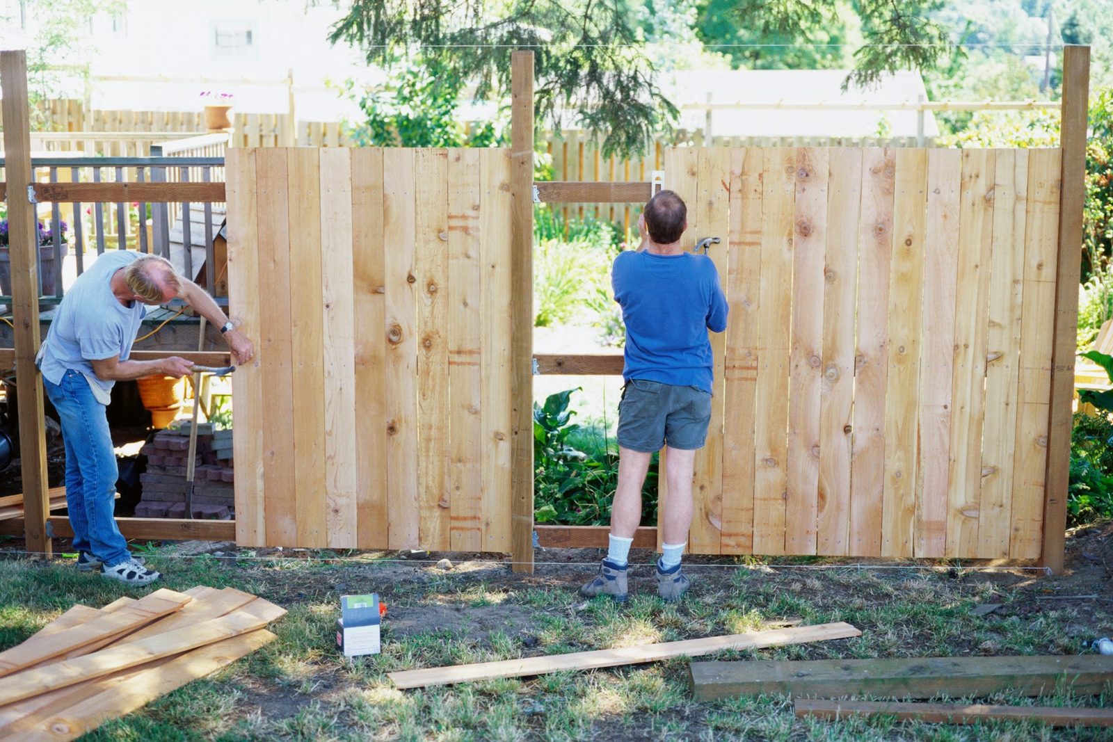 Two men are constructing a wooden fence in a backyard, using tools on a sunny day surrounded by greenery and a wooden deck.