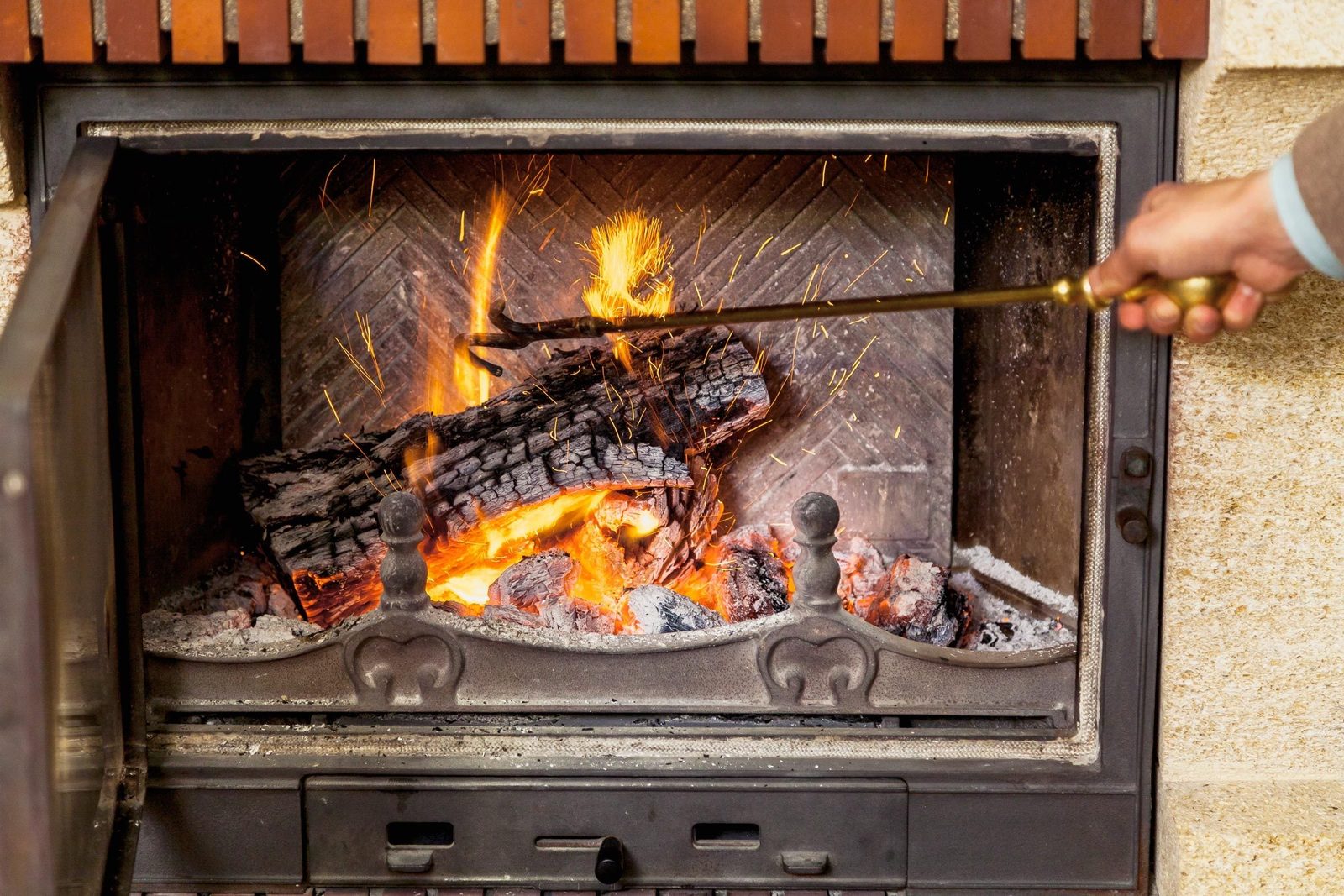 A hand uses a metal poker to adjust burning logs in a fireplace, sending sparks flying against a backdrop of warm orange flames and dark ash