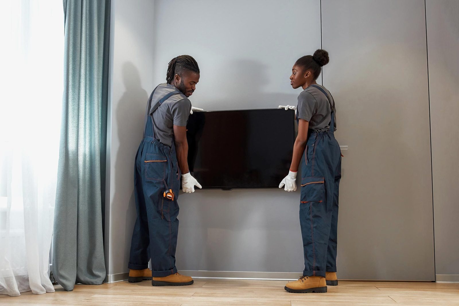 Two individuals lift a television together, adjusting its position against a light-colored wall in a room with curtains and wooden flooring.