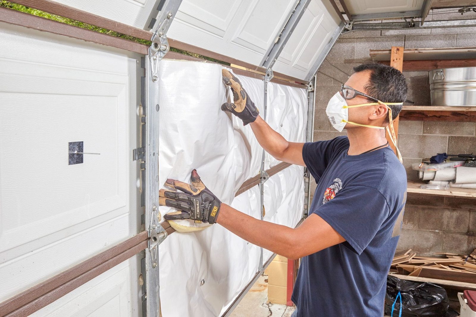 A person wearing gloves and a mask repairs a garage door by applying a white material, surrounded by tools and items in a cluttered garage.